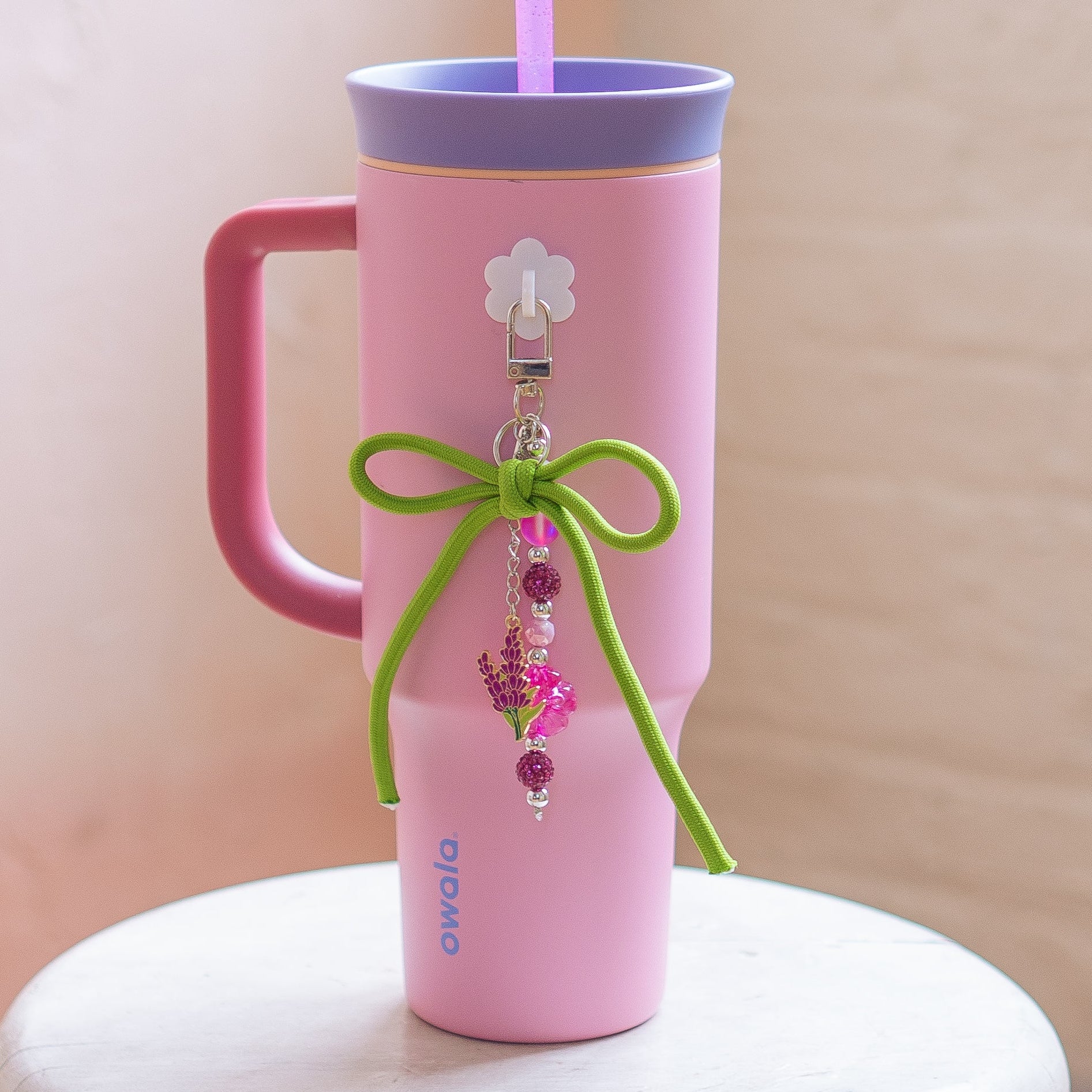 Pink tumbler with a purple lid and handle, decorated with a green bow and pink beads, on a white surface.